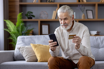 Gray-haired scared man uses credit card through mobile app, holds phone, sits on sofa at home