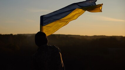 Fototapeta premium Young man in military uniform waving flag of Ukraine against beautiful sunset at background. Male ukrainian army soldier lifted national banner at countryside. Victory against russian aggression