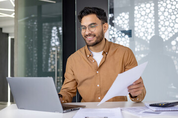 A smiling young Indian man is working in the office with a laptop and documents, holding papers and talking on a video call with clients and partners