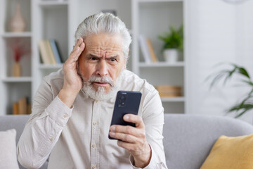 Close-up photo of a shocked senior man in a white shirt at home looking worriedly at the phone screen while holding his head