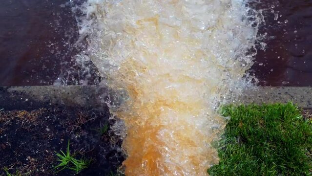 A low, top down shot of a white fire hydrant spraying rusty water onto the street on a sunny day. Filmed in slow motion, the camera tilted downward, dolly out over the rusted spray of water.