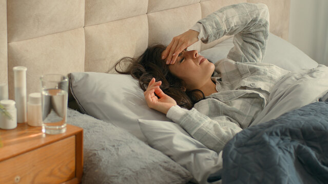 Unhappy woman lying on bed holding head bedroom home girl female headache ill migraine sick rubbing exhaustion healthcare painful suffering unwell upset pressure cephalalgia dizzy temperature disease