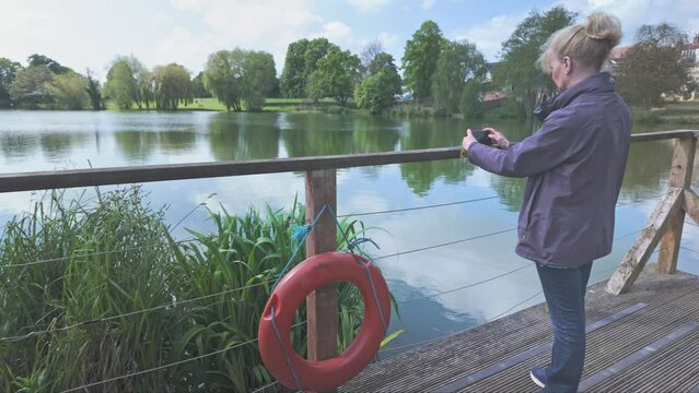Female Tourist takes snapshot of Diss park lake from boardwalk with lifebouy ring