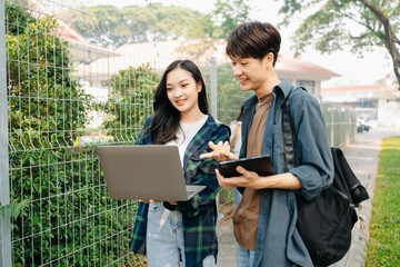 Two smart young Asian college students focusing on his school project, looking at laptop and tablet, discussing and working together