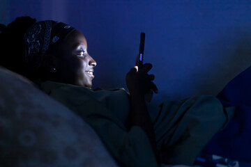 Young African Woman in Dark Room Illuminated by Mobile Phone Light, Highlighting Digital Addiction