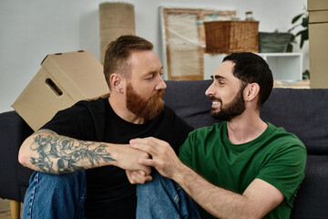 Two men, part of a gay couple, sit happily atop a couch in their new home, amidst moving boxes and...