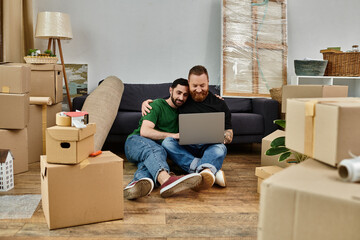 A man and a woman sit on the floor surrounded by boxes, focused on a laptop screen together.
