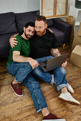Gay couple in love sits on floor, surrounded by boxes, using laptop in their new home.