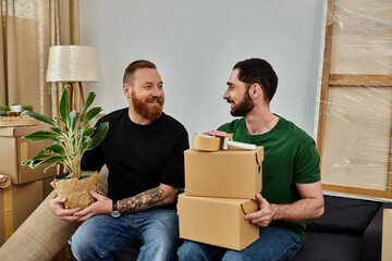 A gay couple in love, holding boxes and a plant, prepares for a new life in their new home filled...