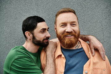 Two men, a loving gay couple, stand side by side wearing casual attire, sharing a tender embrace against a grey wall.