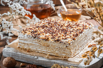 Layered honey cake with sour cream and nuts on light background with cup of tea and flowers. Sweets, dessert and pastry, top view, close up