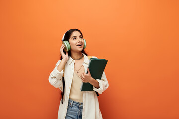 Young indian woman stylishly poses with headphones and folder against bright orange backdrop.