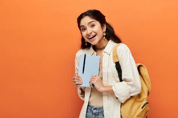 Young indian woman confidently holds notebook on orange background.