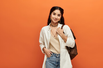 Stylish young indian woman posing confidently in white shirt and jeans against bold orange backdrop.