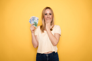 Beautiful young woman in casual clothes joyfully holding in her hand a lot of dollars on a yellow...