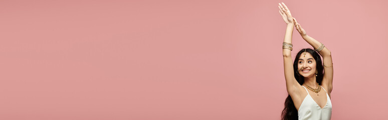 A young indian woman enthusiastically raises her arms on a vibrant pink background.