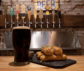 closeup view of a glass of black beer with fried mozzarella sticks on the wooden counter. The beer taps in the background. 