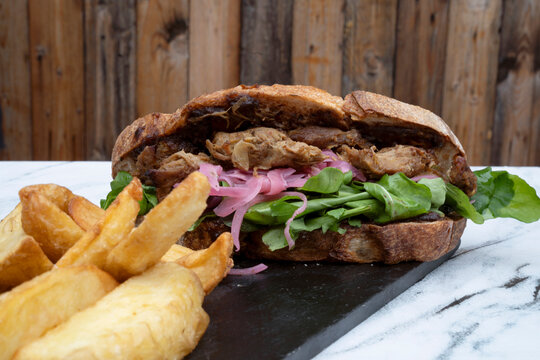 Fast food. Sandwich. Closeup view of a pork meat bondiola sandwich with lettuce, onion pickles and fried potatoes in a black dish on the pub white marble table.