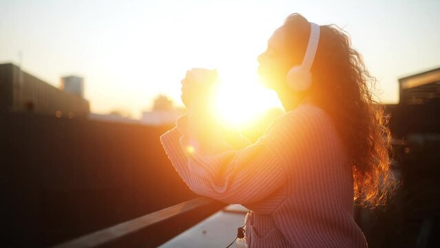 A Young, Inspired Woman Wearing Headphones is Drinking Coffee or Tea while Standing on a Terrace and Admiring the City's View. She is Listening to Her Favorite Playlist. It's a Beautiful Calm Morning