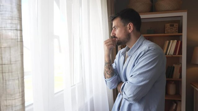 Young sad and depressed man feeling regret and anxiety, standing near window looking through. Mental burnout after hard work and hours of insomnia. Relationship difficulties desperation and depression