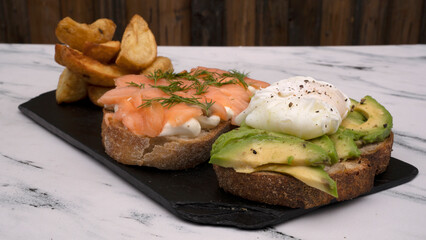 Healthy cuisine. Gourmet sandwich. Top view of a smoked salmon, cream cheese, sliced avocado and poached eggs sandwich with fried potatoes on a black dish on the white marble table.