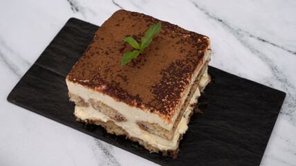 Italian food. Sweet desserts. Closeup view of a square slice of Italian dish tiramisu. Coffee flavor, mascarpone cheese, and chocolate cake in a black dish with a wooden background.