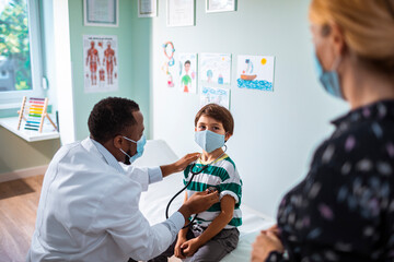 Pediatrician examining young boy's heart with stethoscope in clinic while mother watches