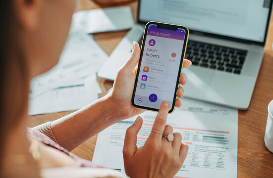 Woman using banking app on smartphone with laptop and bills on desk