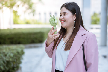 a young woman standing outdoors, smiling while holding a small, handheld fan. She appears to be enjoying the breeze from the fan.  © Anucha