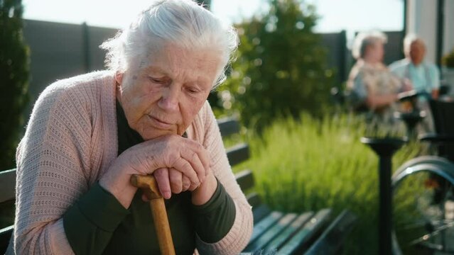 Lonely worried anxiety woman in the 90s sits on a bench in the yard of a nursing home pensive and nostalgic, feel weak hopeless