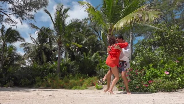 Cheerful couple in background of scenic tropic landscape performs lambdas barefoot on soft sand, moves young man pulls girl off ground, bends her in back and then presses her against him