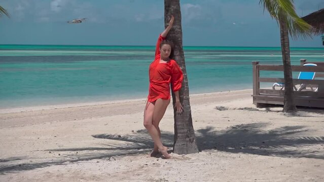 Dancing girl in red dress at ocean shore leans on palm tree posing, quadcopter flies over water doing shooting aerial side view, girl looks in drone direction, lifts up trained hip and stretches