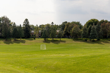 Vast green grassy field - partly cloudy sky - scattered trees - soft shadows - white goal post and residential area in background. Taken in Toronto, Canada.