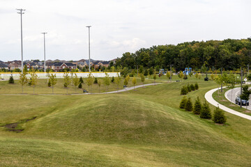 Suburban park with green rolling hills - paved walkways and young trees - playground equipment - residential houses background - tall light poles and partly cloudy sky. Taken in Toronto, Canada.