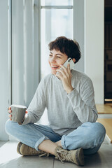Cheerful young woman talking on the phone and drinking coffee in the morning.