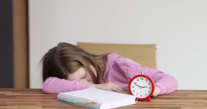 Little girl falling asleep at table with red alarm clock and textbook at home 4k movie slow motion. Lack of sleep in children concept