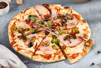 Artisanal pizza topped with ham, mushrooms, olives, and fresh garnishes, served on a textured gray background, showcasing a hearty and delicious meal