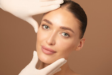 Cosmetic surgery concept. Doctor examining young woman's face over beige studio background. Beautiful lady during visit to cosmetologist cabinet
