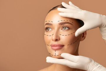 Portrait of young woman and plastic surgeon making marks on female face over beige studio...