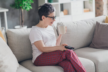 Photo of pretty retired woman watch tv drink tea sit sofa wear white outfit home flat rest relax spend free spare time living room indoor