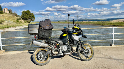 Obraz premium This image shows a black and yellow adventurer's motorcycle parked next to a river, fully equipped with travel gear including a large backpack and a storage box securely attached to the back