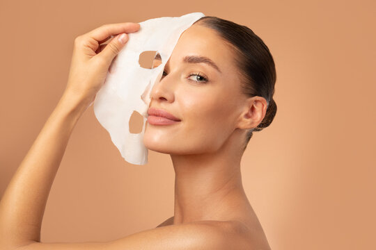 Beautiful European lady removing moisturizing white fabric sheet mask from her face after using it, looking at camera, posing on beige background