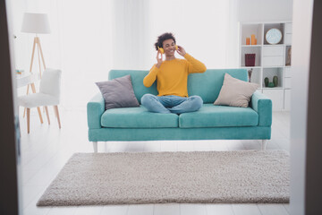 Full length portrait of nice young man sit sofa headphones wear pullover white interior flat indoors