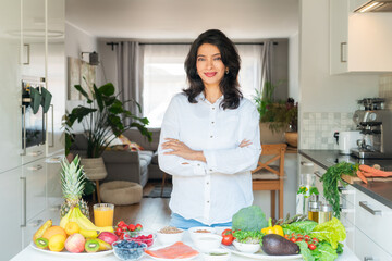 Confident smiling middle aged female Indian Nutritionist expert arms crossed in front of kitchen table full of fruits and vegetables. Healthy diet habits and wellness lifestyle, weight loss program.