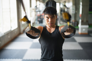 A focused man in a black tank top uses suspension training straps in a gym. His intense expression...