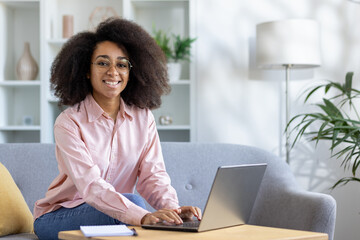 A woman happily works on her laptop in a home office with natural light and plants, creating a cozy...