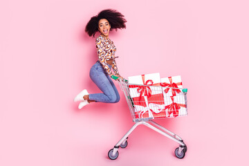 Full length photo of charming girly woman wear print shirt jumping walking supermarket trolley buying presents isolated pink color background