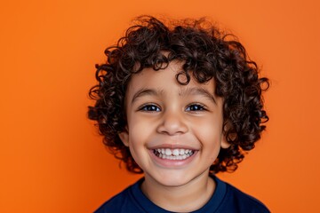 young boy with dark brown curly hair and light brown skin, wearing dark blue t-shirt showing a toothy grin against a bright orange background