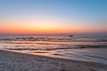 A serene sunset over a calm sea with gentle waves lapping at a pebbled beach. A small boat is silhouetted against the vibrant hues of the sky, creating a tranquil and picturesque scene perfect for con