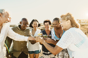 Happy multiracial senior friends having fun drinking and toasting mojitos on the beach during sunset time - Elderly people enjoying summer holidays
